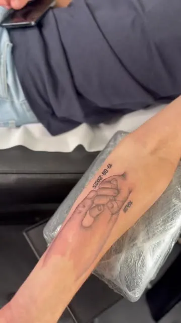 Deux mains qui se tiennent, tatouées en noir et gris sur un avant-bras, avec une date.