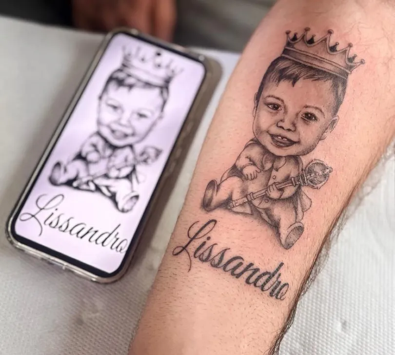 Portrait d'un petit garçon avec une couronne et un sceptre tatoué sur un avant-bras, au-dessus du prénom Lissandro, à côté du modèle affiché sur un téléphone.